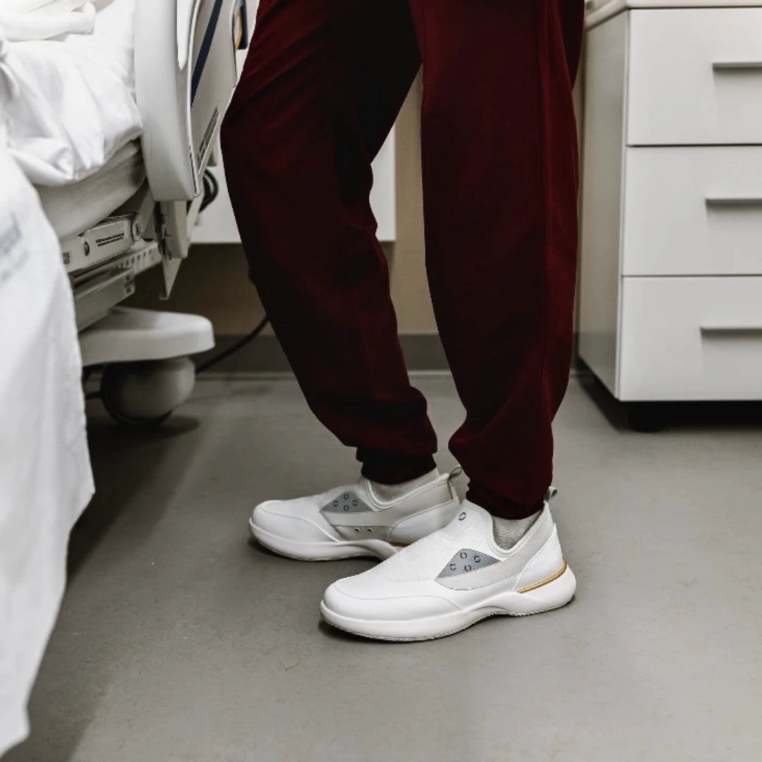 Nurse in scrubs wearing white Snogs Genesis Day Shift sneakers beside a hospital bed